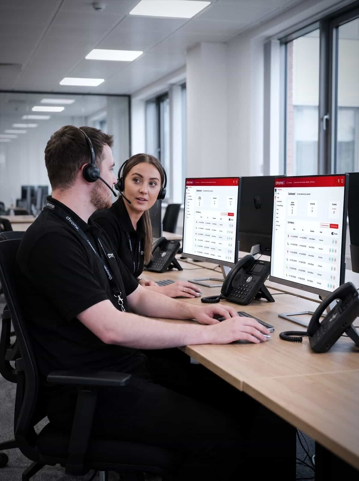 Two Chrome Telecom colleagues at adjacent desks with headsets and VoIP handsets, talking between calls