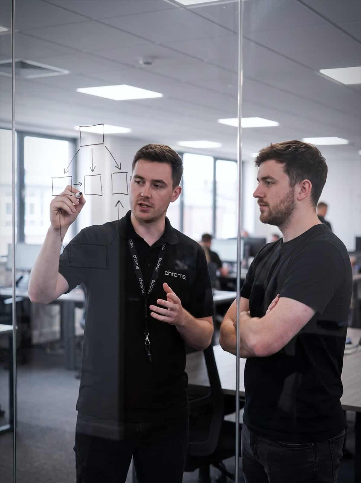 Two Chrome Telecom colleagues sketch a systems plan on a glass wall in a meeting room