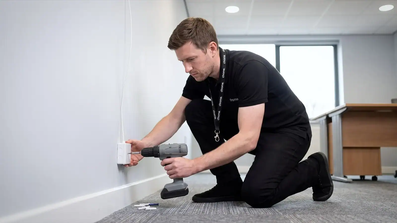 A Chrome Telecom engineer kneeling by an office wall, fixing a small white fibre optic termination box above the skirting board with a cordless drill