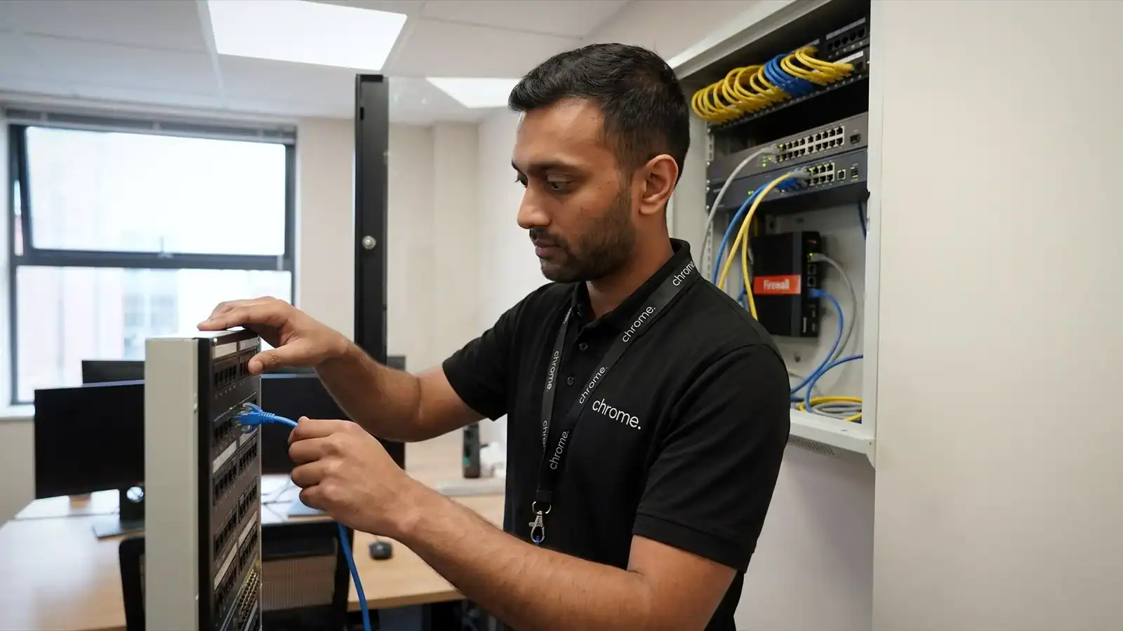 A Chrome Telecom network engineer patching a cable into a switch in a wall-mounted comms cabinet in a small UK office