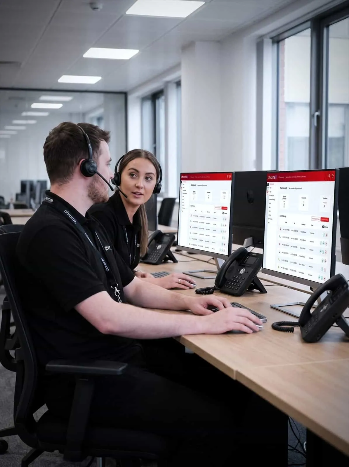 Two Chrome Telecom colleagues at adjacent desks with headsets and VoIP handsets, talking between calls