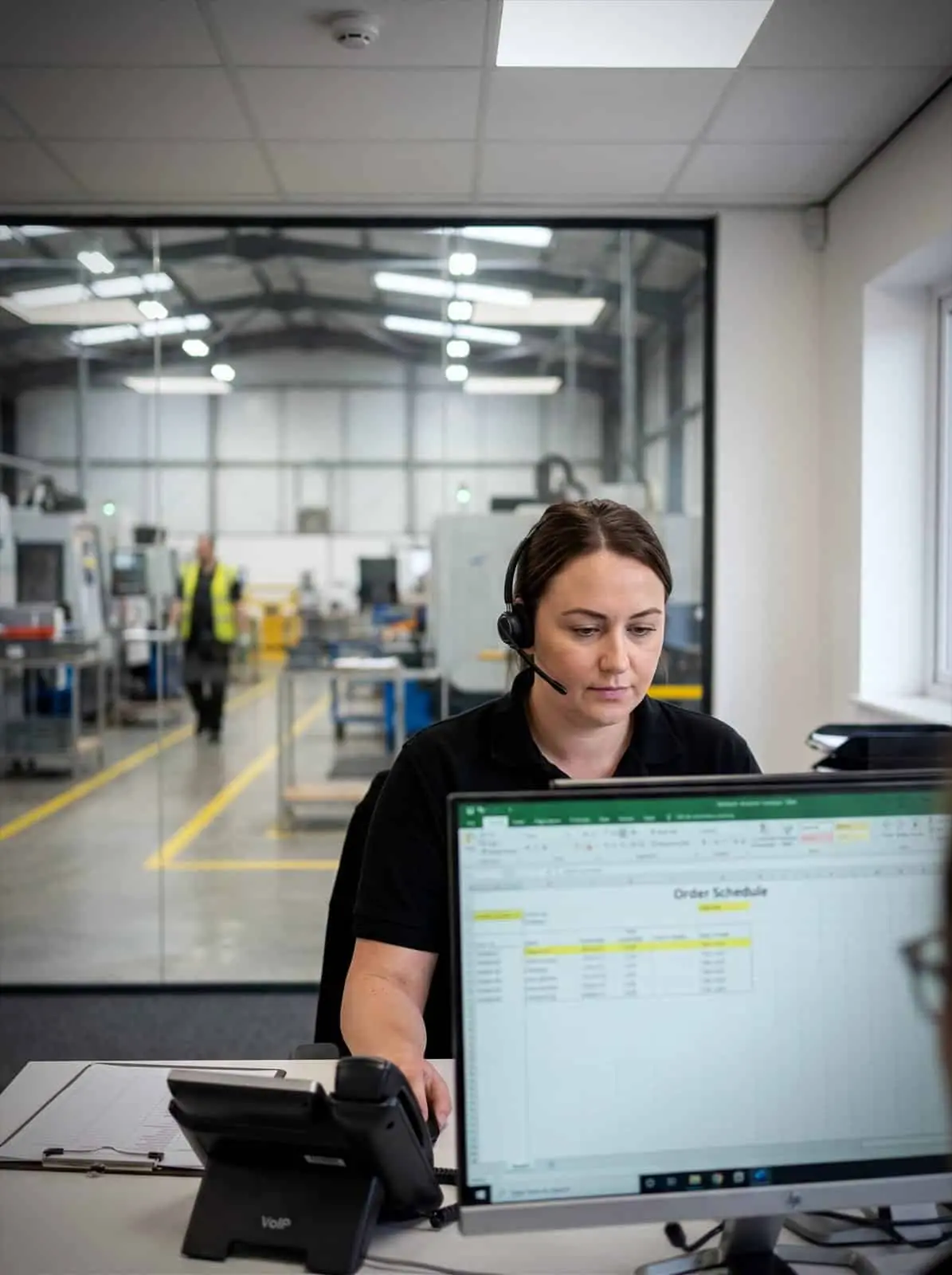 A production coordinator takes a supplier call in an office overlooking the factory floor